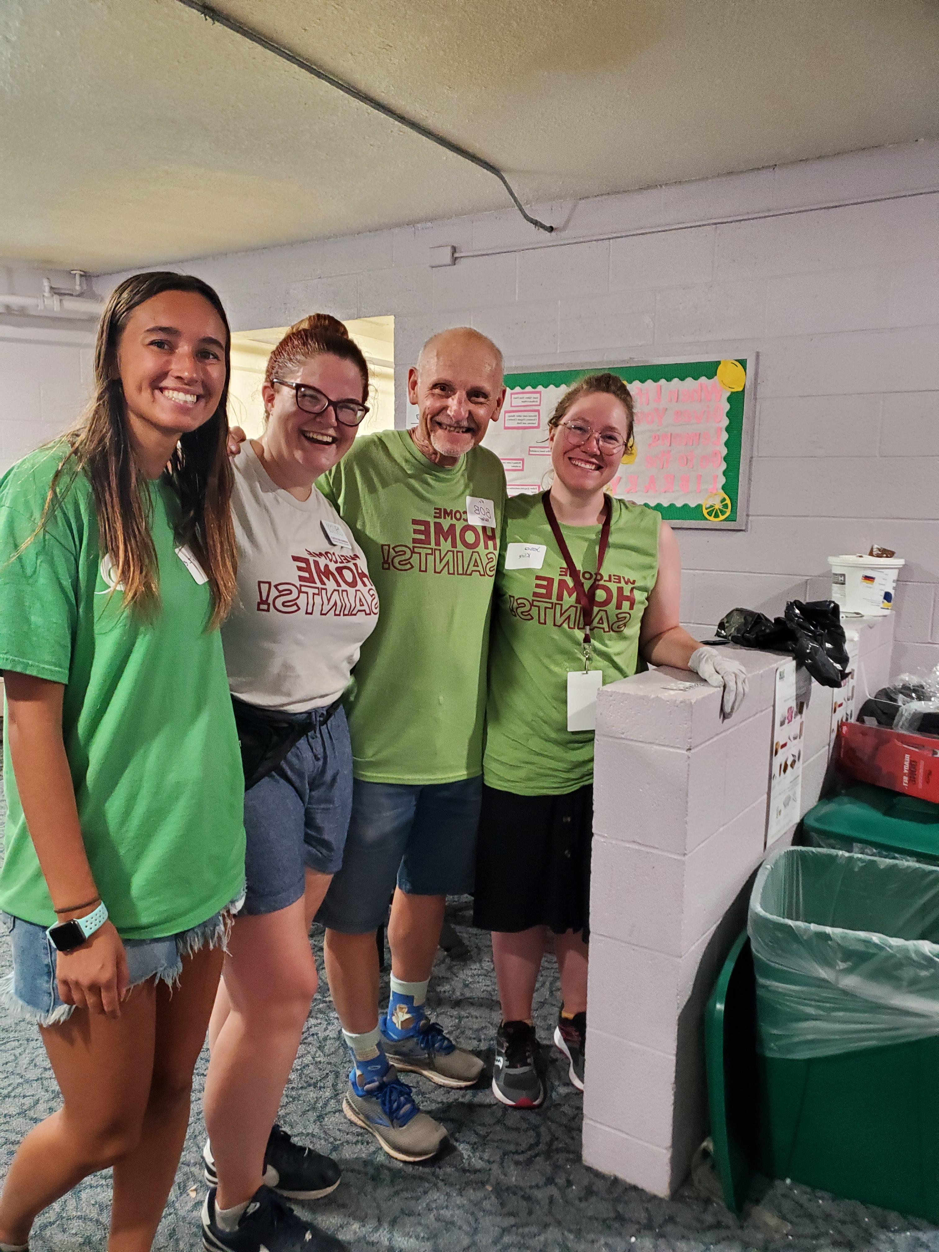 Four people smiling in shorts, sneakers, AQ T shirts next to recycling bins